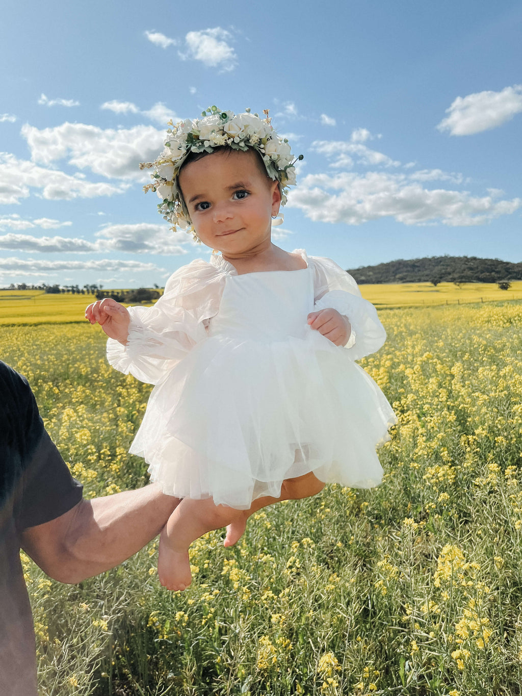 Eva baby flower girl dress is worn by a baby girl. She sits in a flower field holding a flower.
