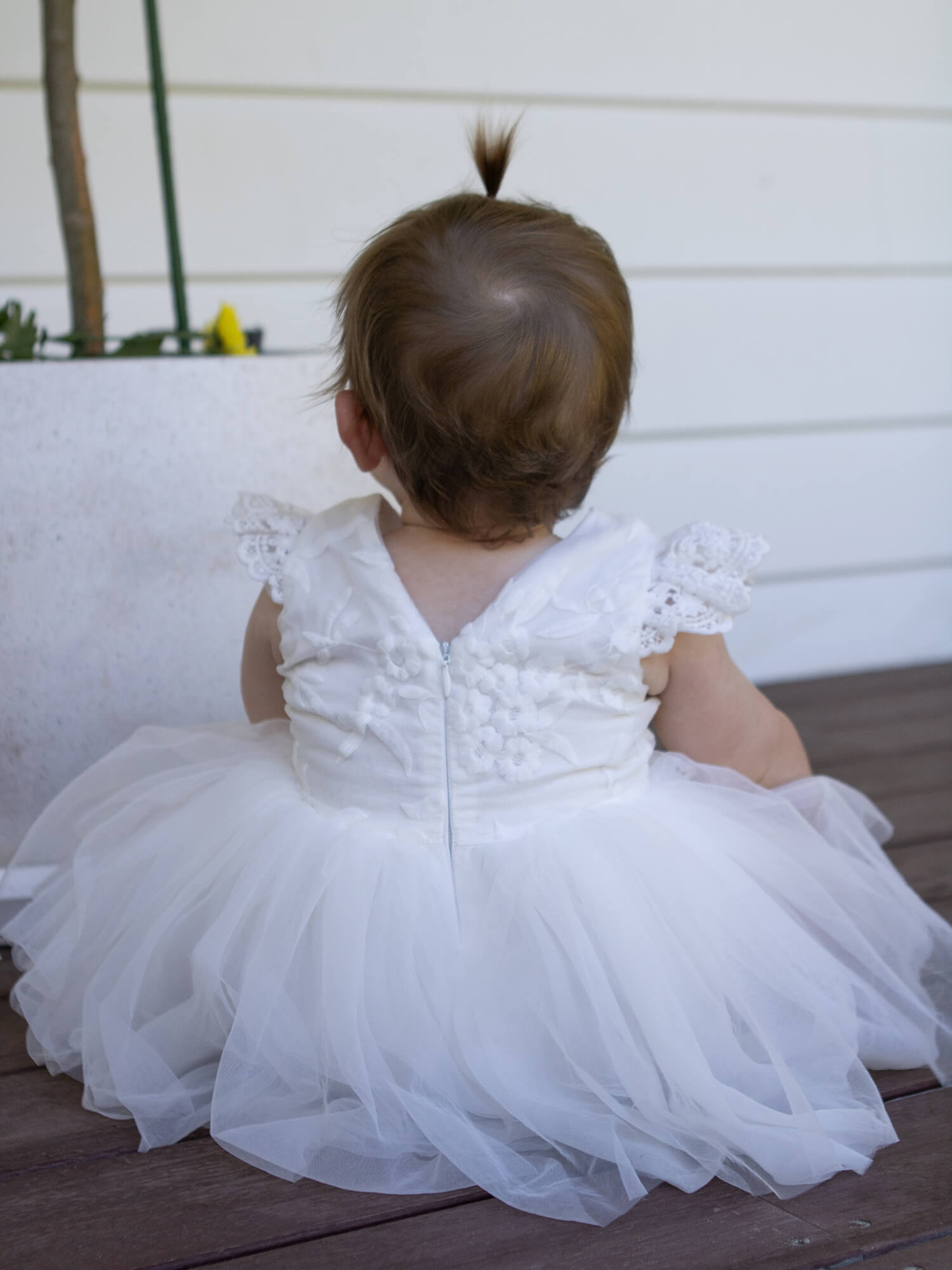 A baby sits in a white lace and tulle baby dress.