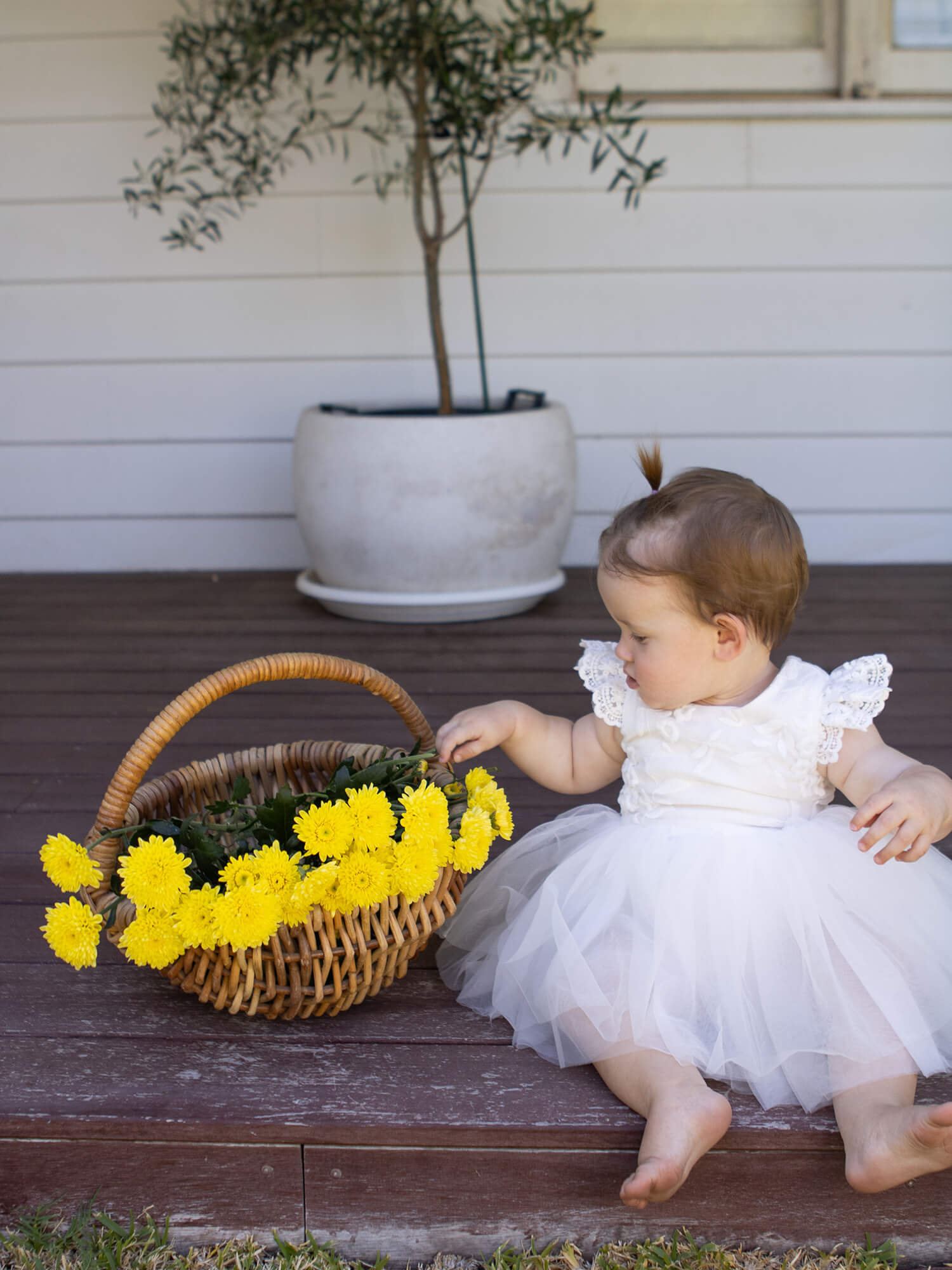 Abigail baby flower girl dress is worn by a baby girl who sits next to a basket of yellow flowers.