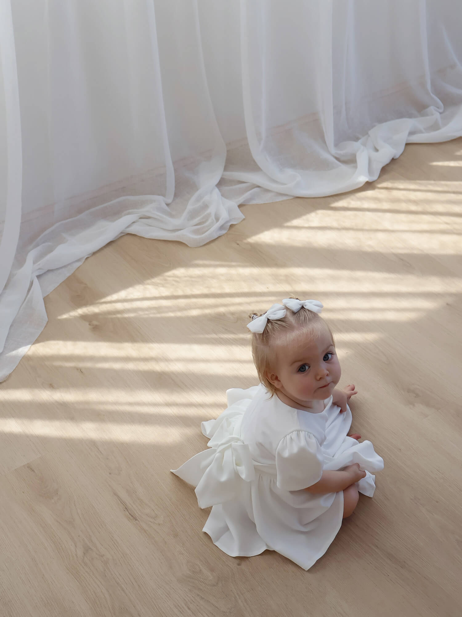 A toddler sits wearing a crepe ivory flower girl dress and matching tulle hair bows.