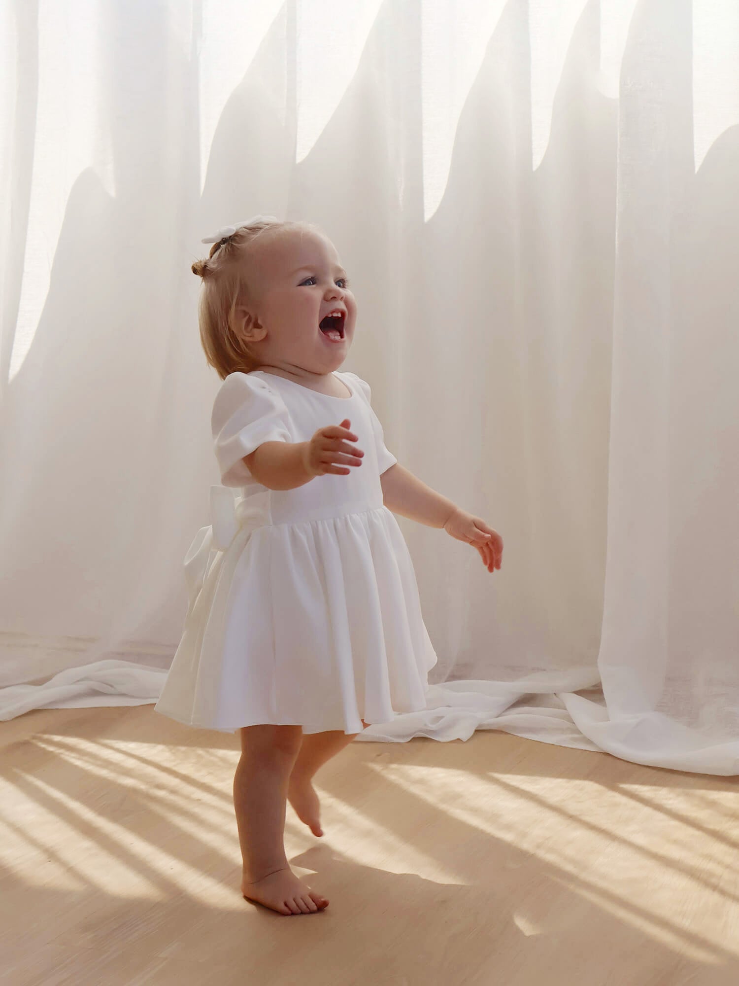 A toddler laughs while wearing an ivory flower girl dress with cap sleeves and a bow at the back.