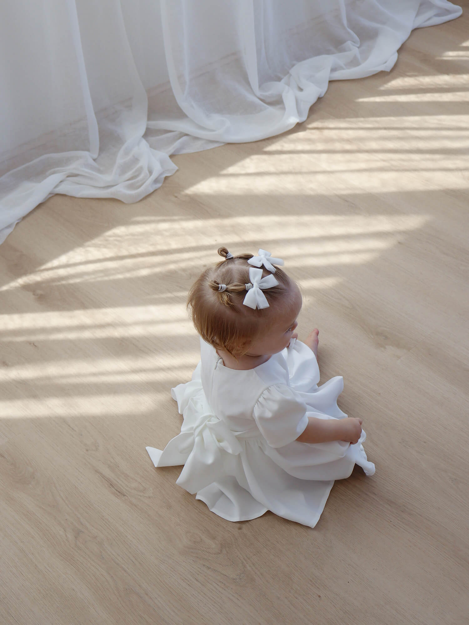A toddler sits wearing tulle bows in her hair and an ivory baby flower girl dress.