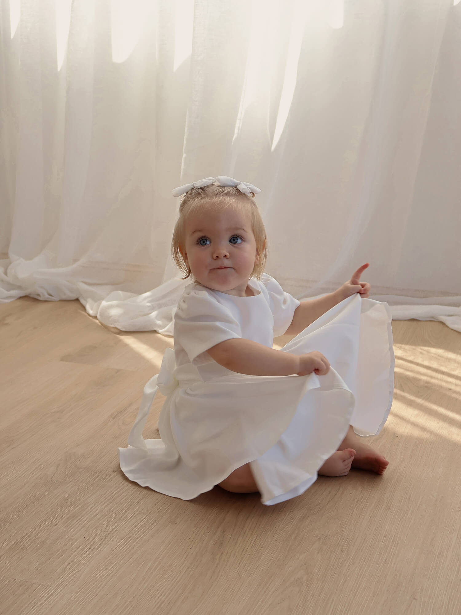 A toddler sits playing with her ivory flower girl dress.