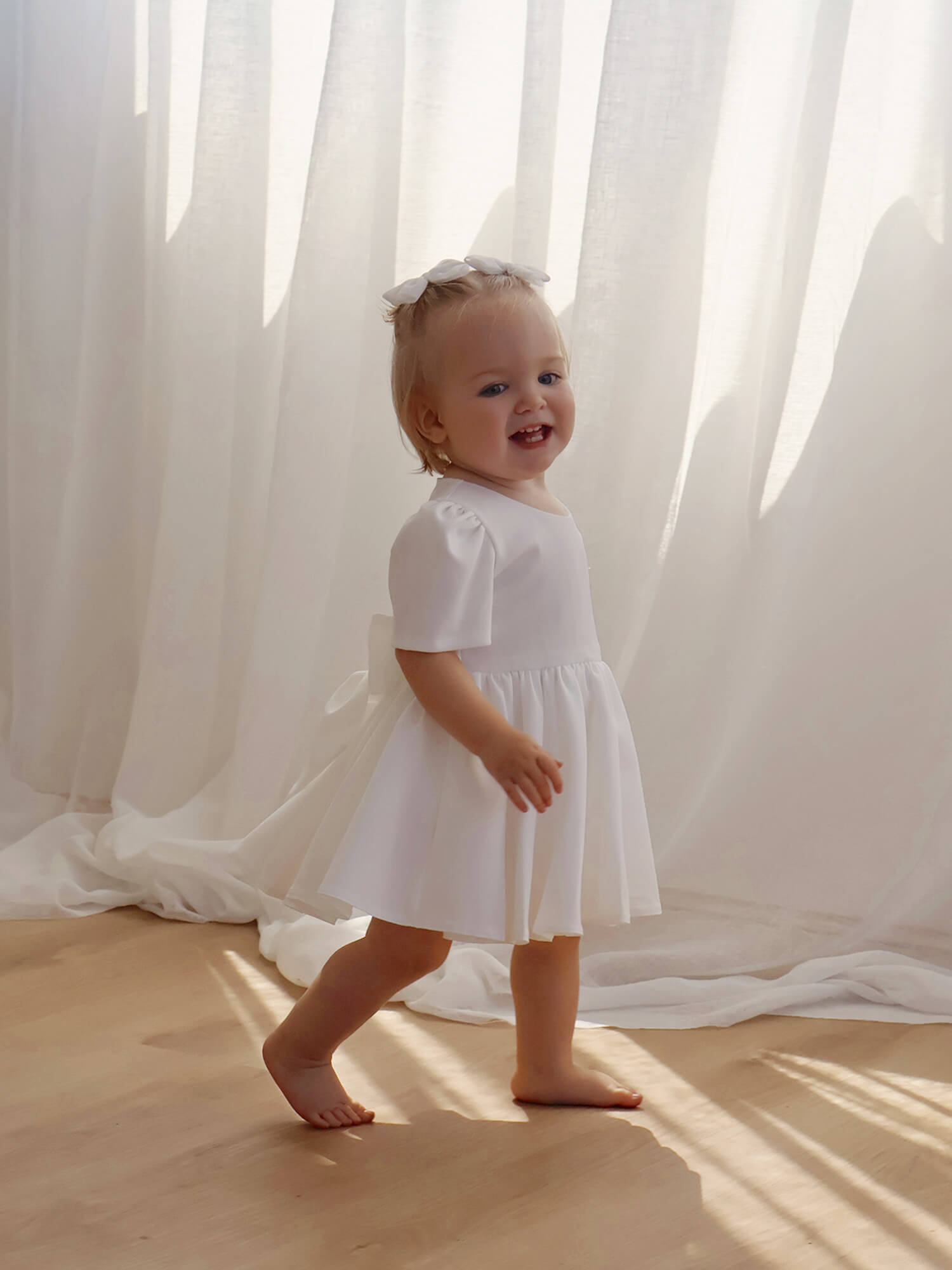 Child in a white flower girl dress standing in front of sheer curtains