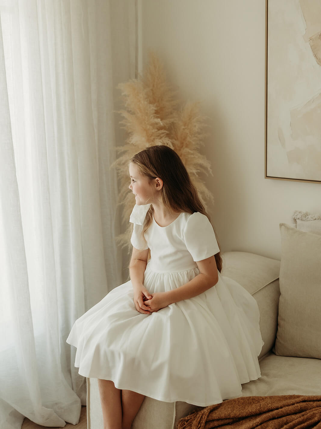 Young girl in a white flower girl dress sitting on a couch in a softly lit room with white curtains.