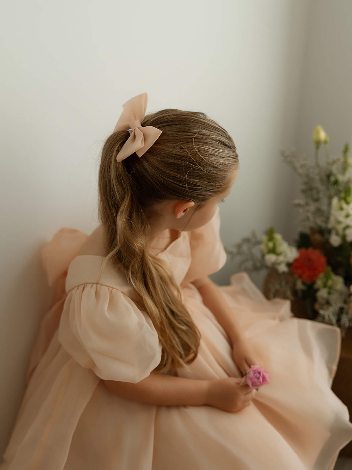 A young girl sits wearing our champagne puff sleeve Cleo flower girl dress and matching champagne organza bow.