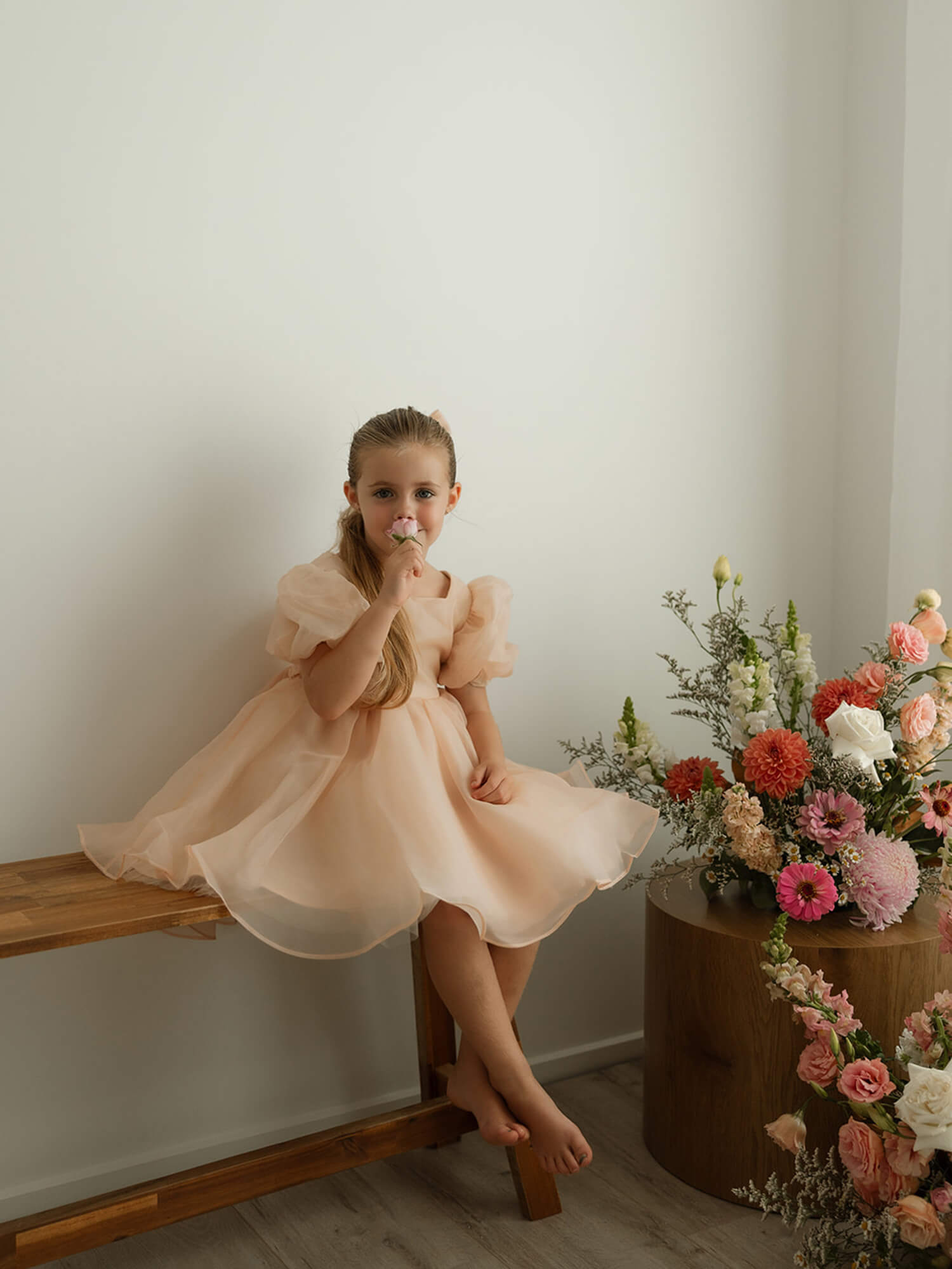 A young girl sits smelling a flower, wearing our Cleo champagne puff sleeve flower girl dress.