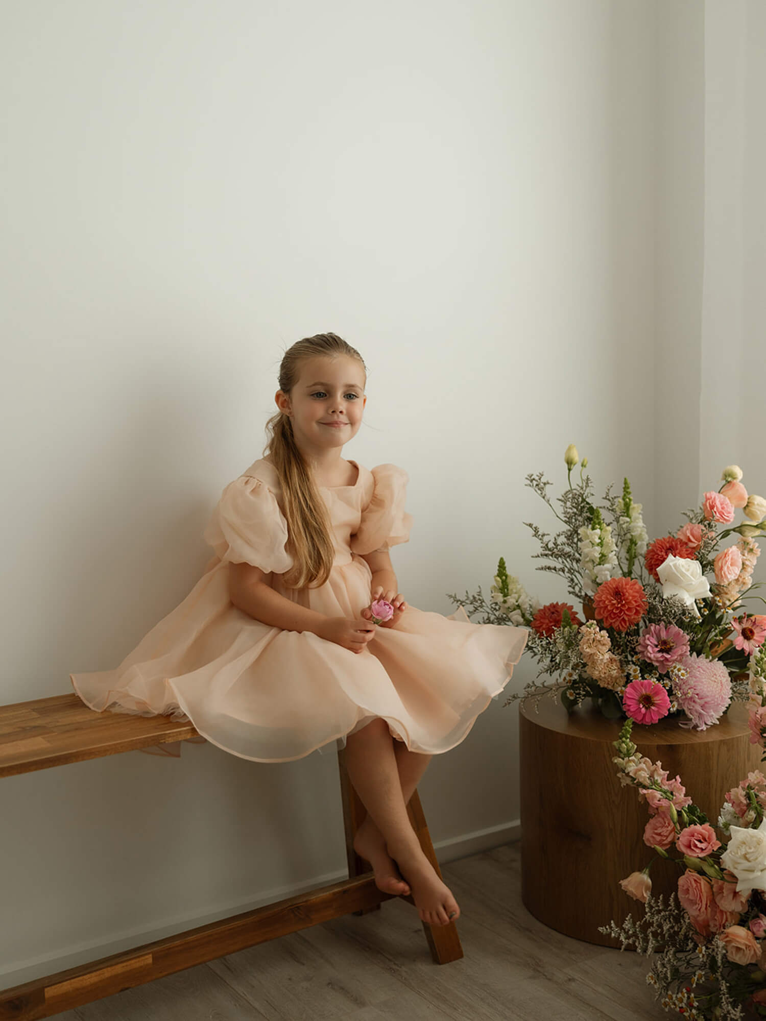 Cleo champagne organza flower girl dress is worn by a young girl, who sits beside flowers.
