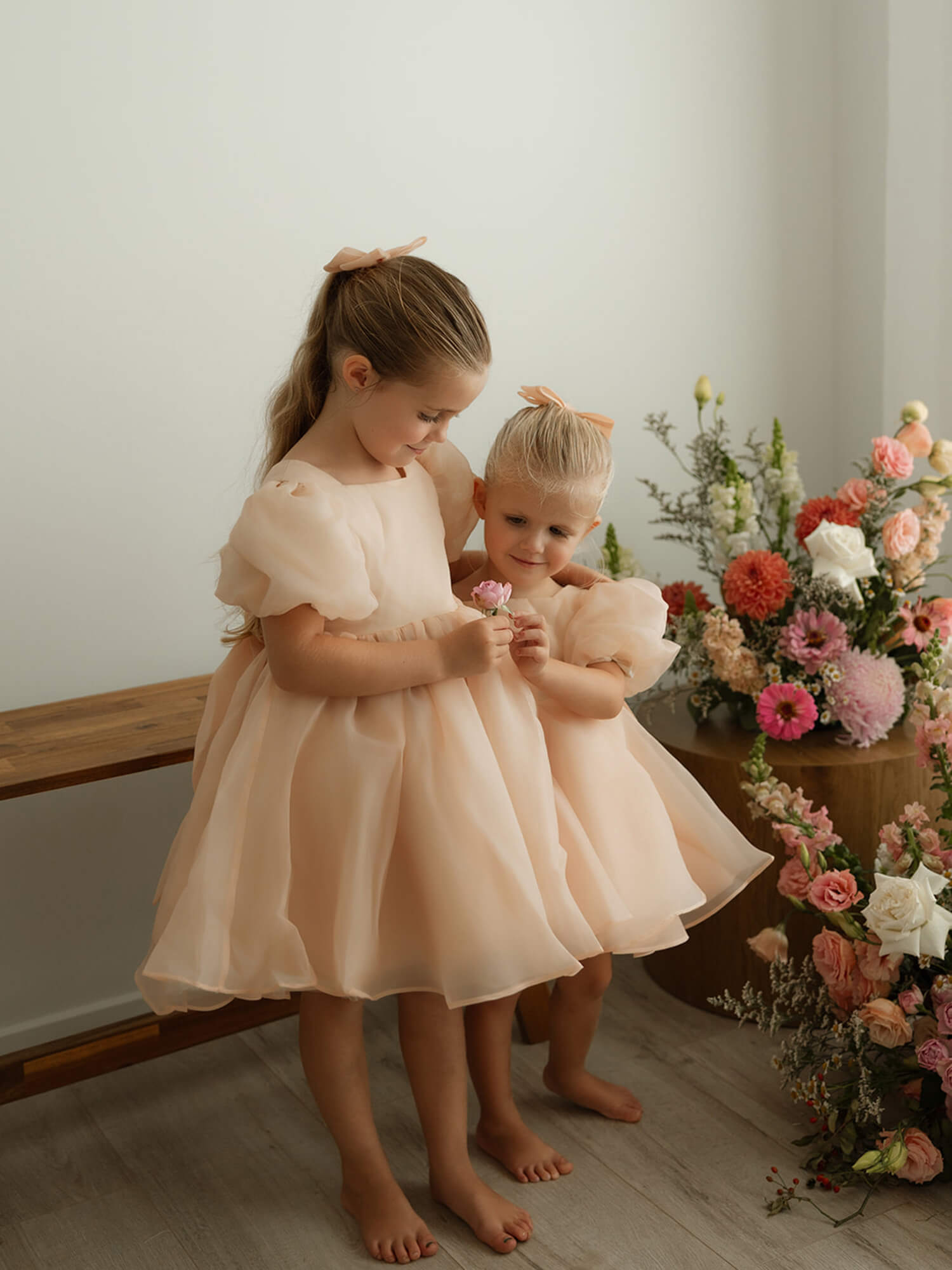 Sisters hug wearing matching Cleo champagne flower girl dresses in champagne organza.