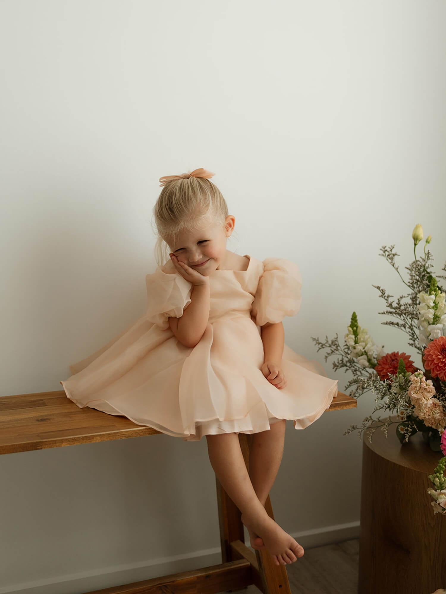 A toddler smiles with her head leaning on her hand, she wears our Cleo champagne baby flower girl dress.