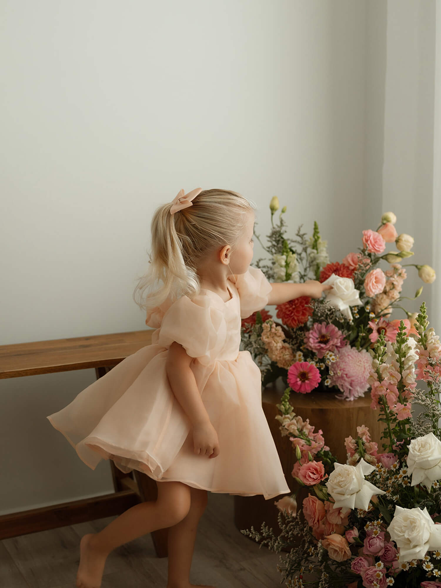 A toddler reaches for a flower, wearing our Cleo champagne organza puff sleeve baby flower girl dress.