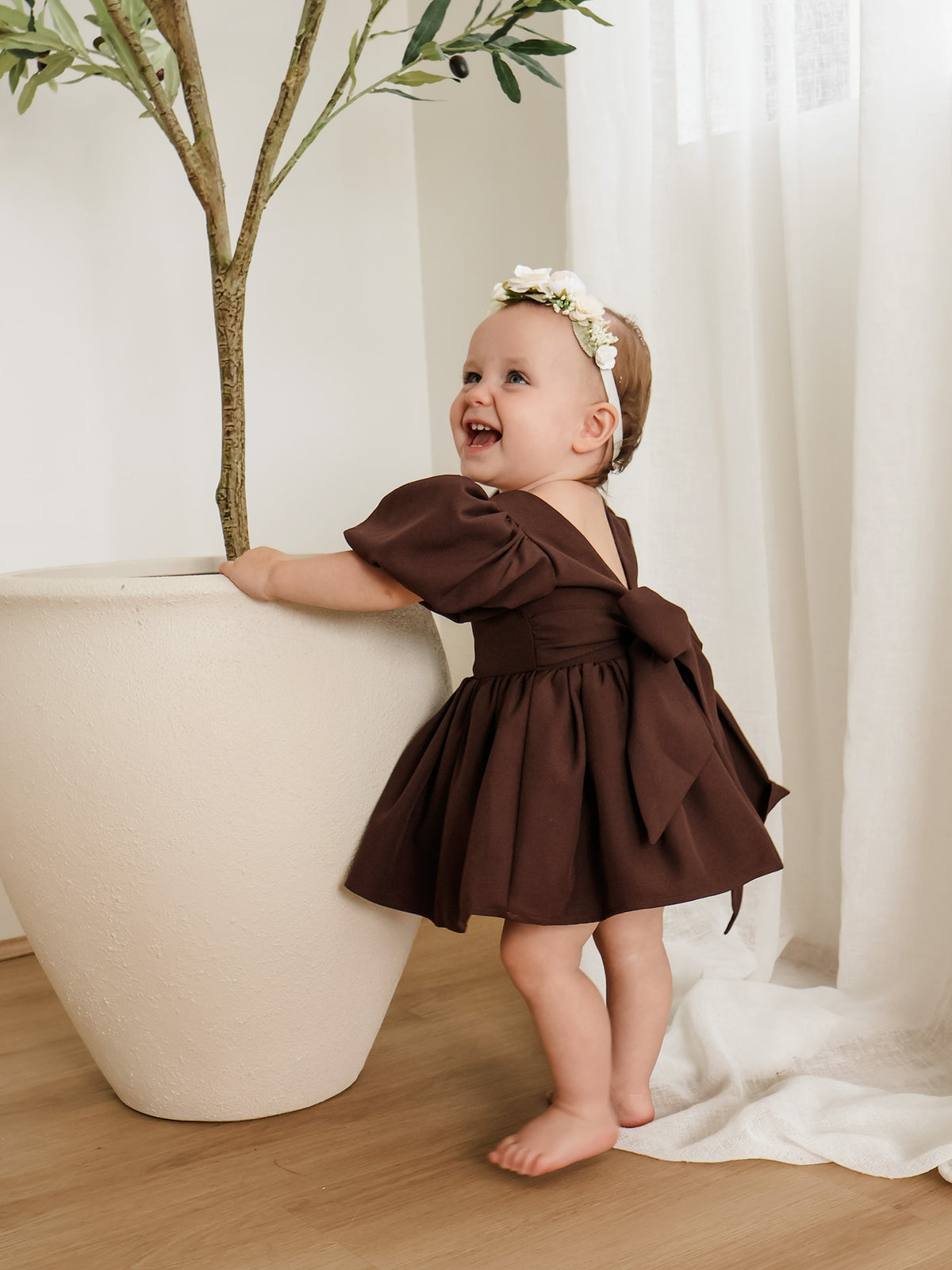 A toddler wears our Cleo chocolate crepe baby flower girl dress and an ivory floral headband.