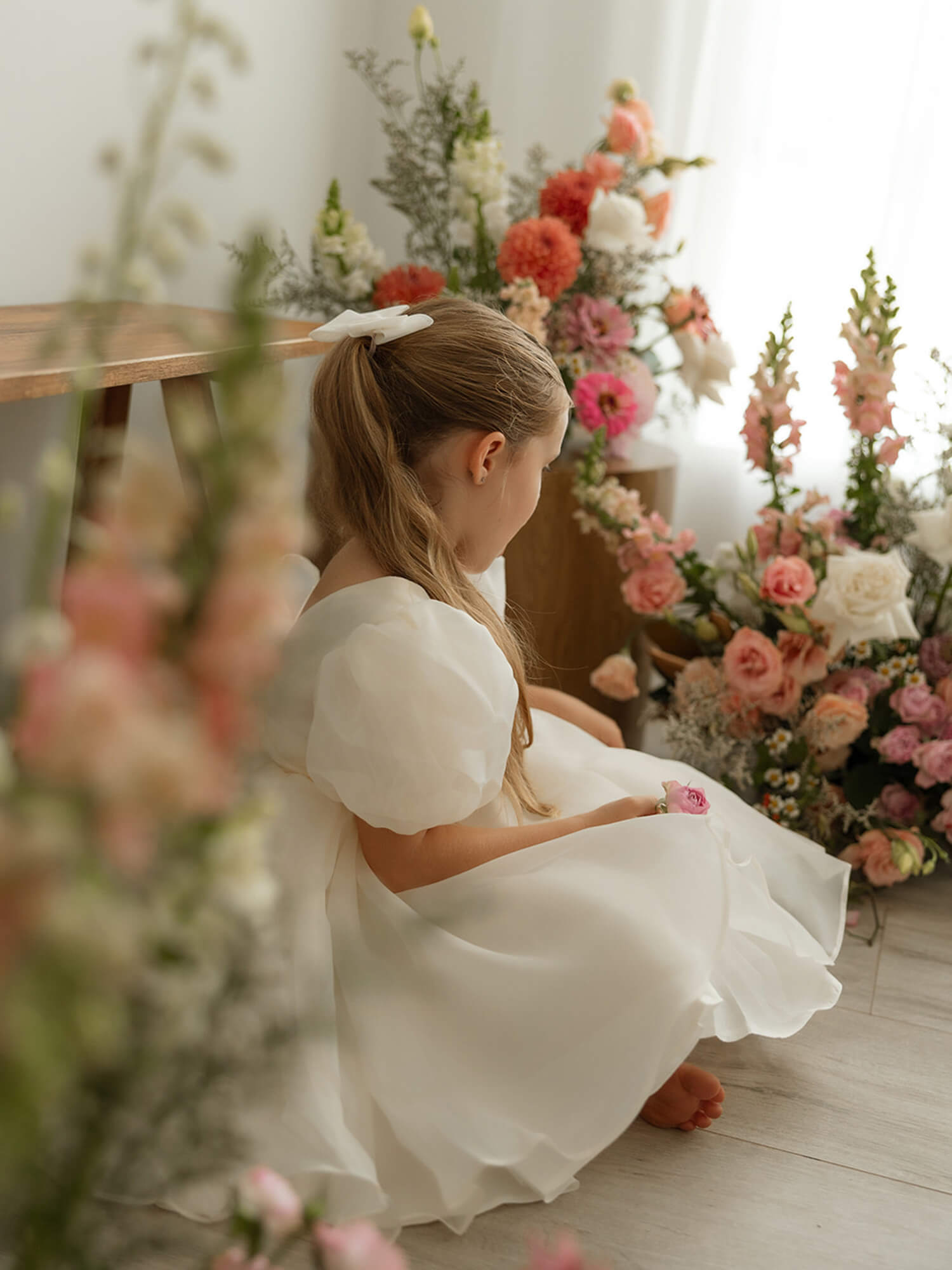 Cream puff sleeved flower girl dress, our Cleo dress in cream organza, is worn by a little girl who is sitting amongst vases of flowers.