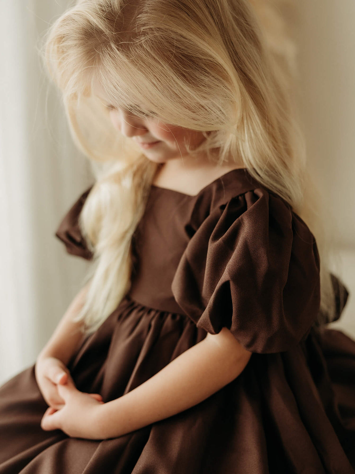A young girl with long blonde hair wearing a dark brown puff-sleeve dress, sitting in a softly lit indoor setting.