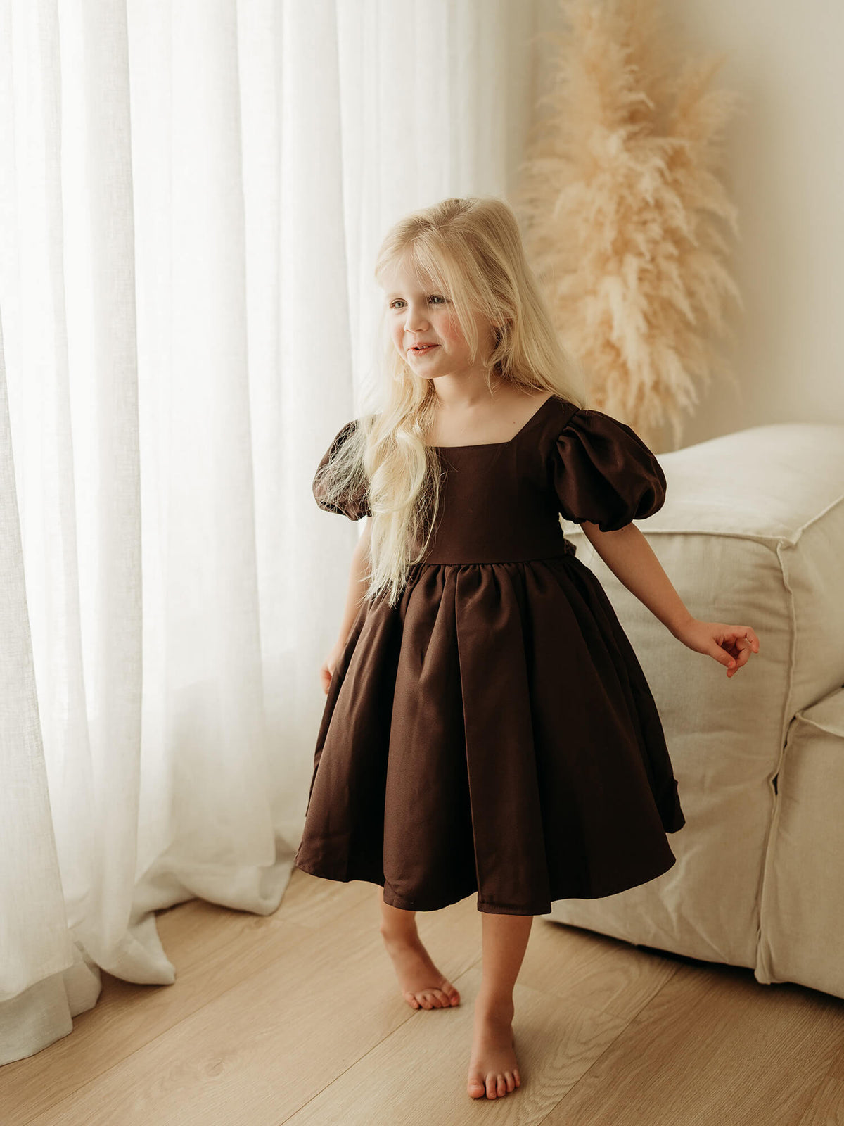 Chocolate brown puff sleeve flower girl dress is worn by a young girl standing near a sheer curtain.