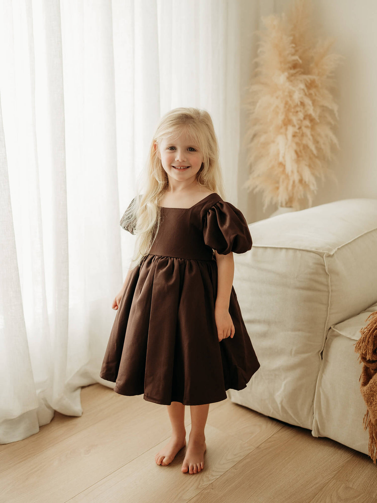 A young girl stands in a bright room wearing a dark chocolate brown girls dress with puff sleeves and a square neckline.