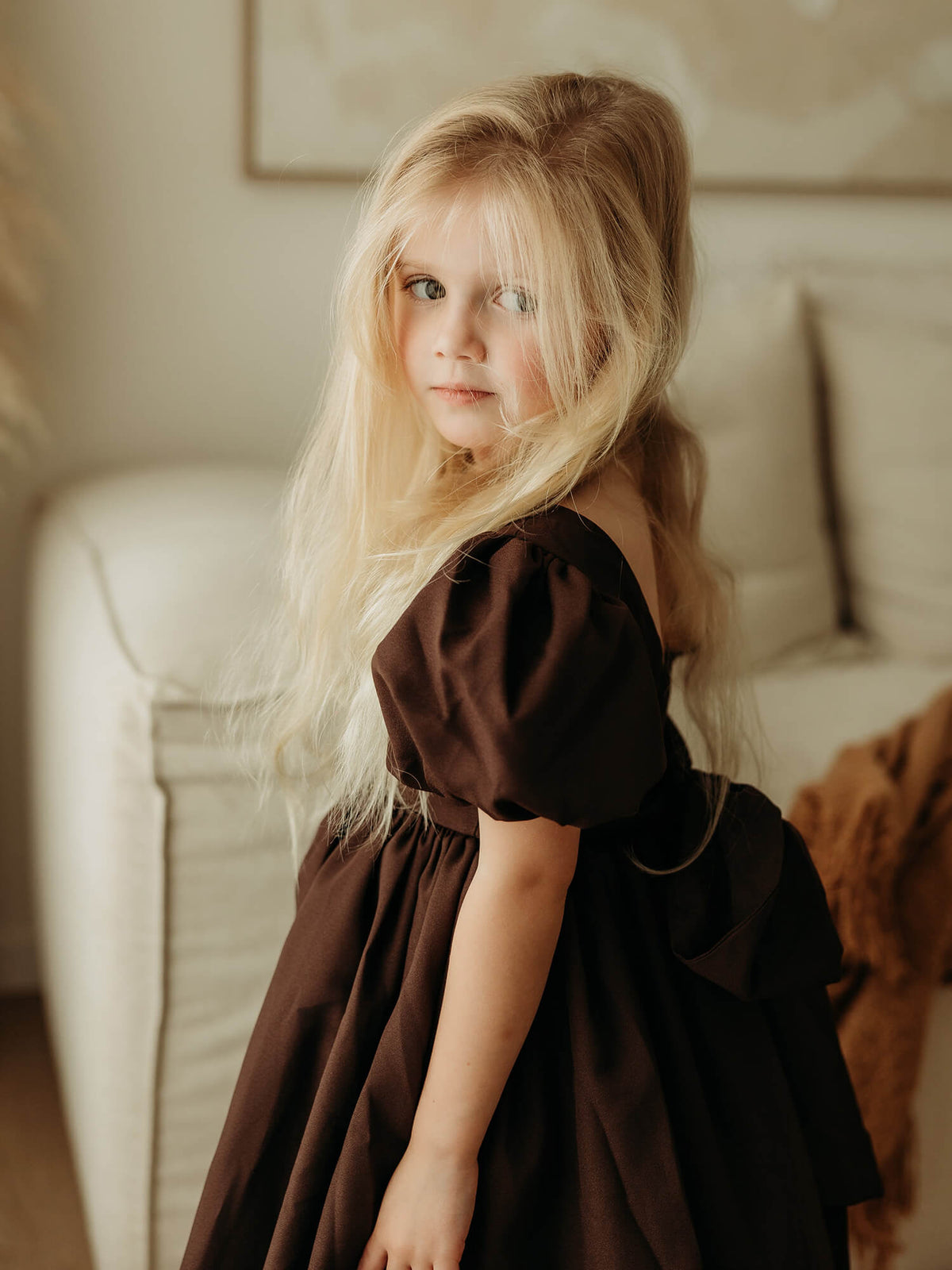 A young girl wearing a chocolate brown puff sleeve flower girl dress, standing in a softly lit indoor setting.