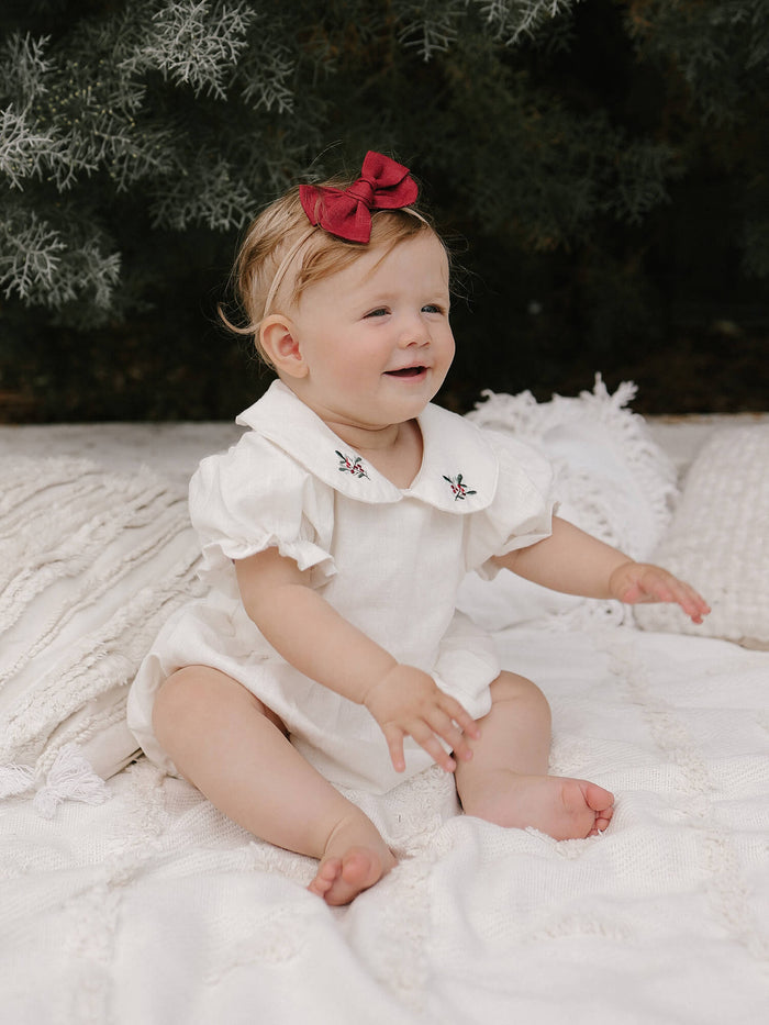 Emma Christmas romper is worn by a baby girl. A perfect Christmas outfit made complete with a crimson bow headband.
