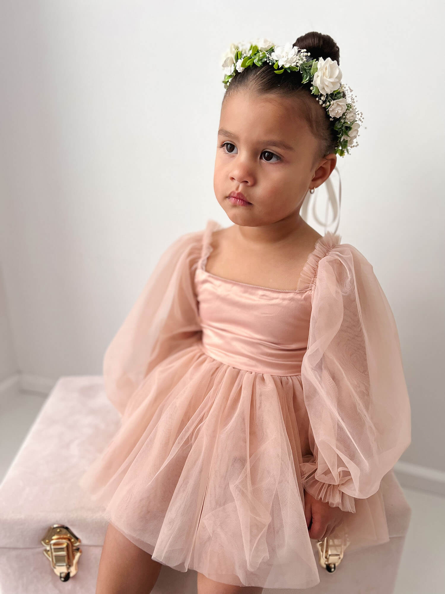 A little girl sits wearing our Eva champagne baby flower girl romper dress and Elsie flower crown in ivory.