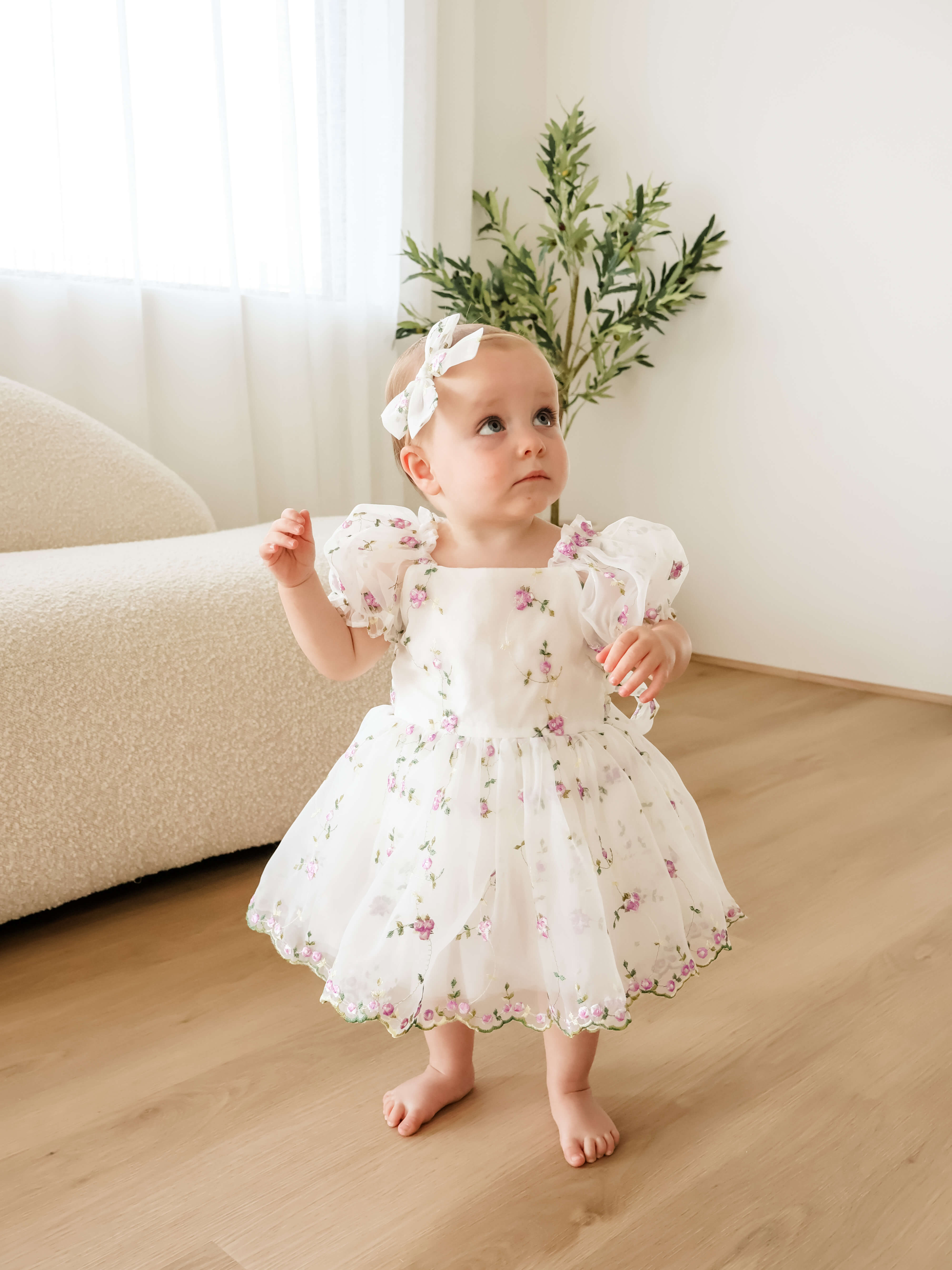 A toddler girl wearing a white puff-sleeve flower girl dress with a lilac floral print, matching headband, and standing barefoot on a light wood floor in a bright room.