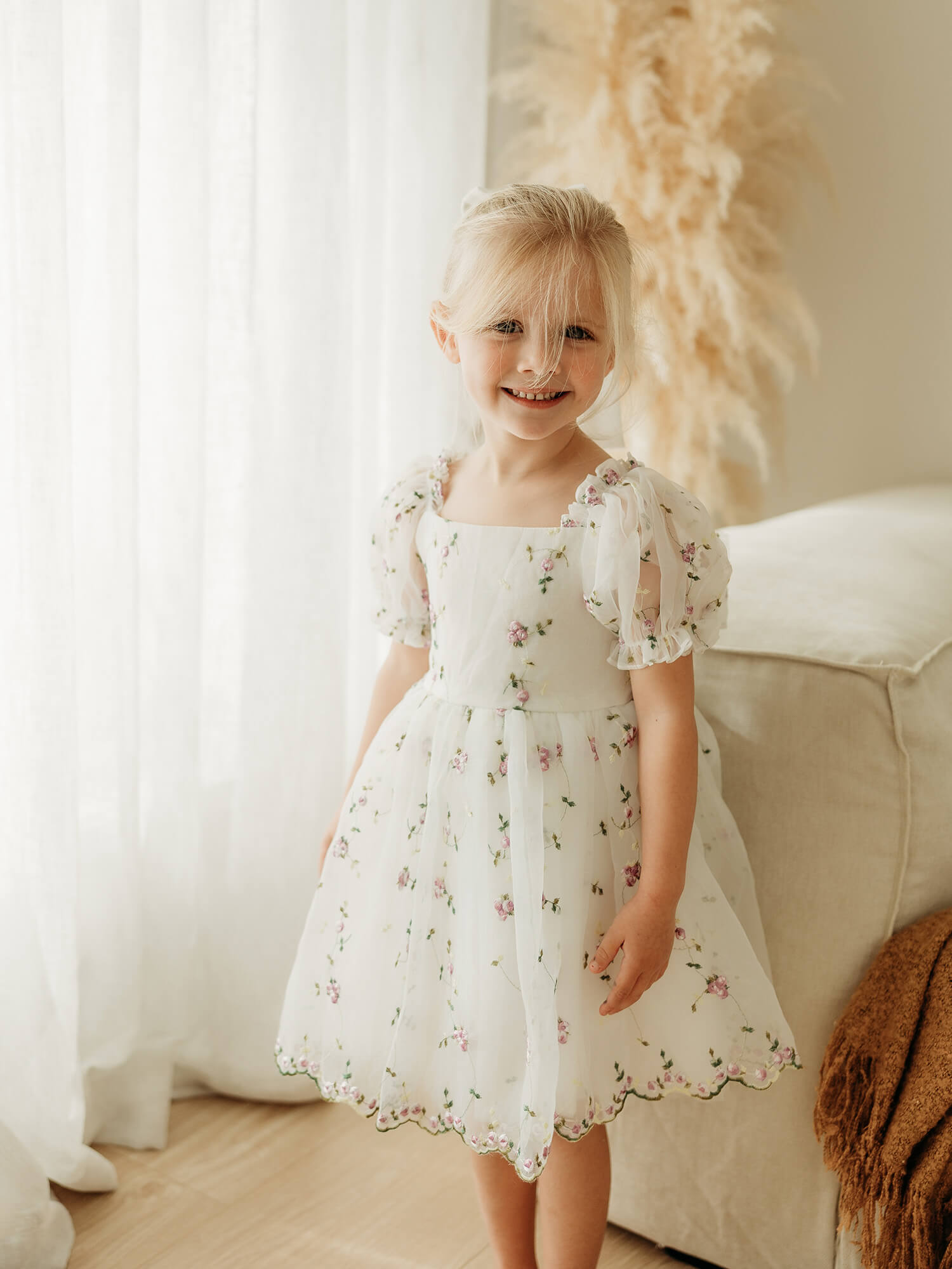 A young blonde girl wearing our Fleur ivory flower girl dress with delicate lilac floral embroidery, standing in a brightly lit room with sheer curtains, a beige sofa, and dried pampas grass.