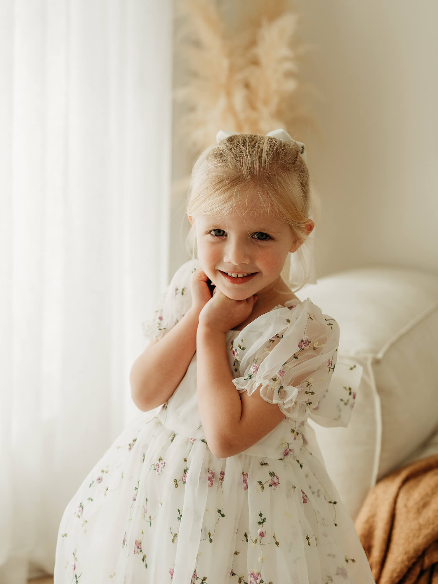 A young blonde girl smiling in a white organza flower girl dress with a delicate lilac floral print, standing in a brightly lit room with soft neutral decor.