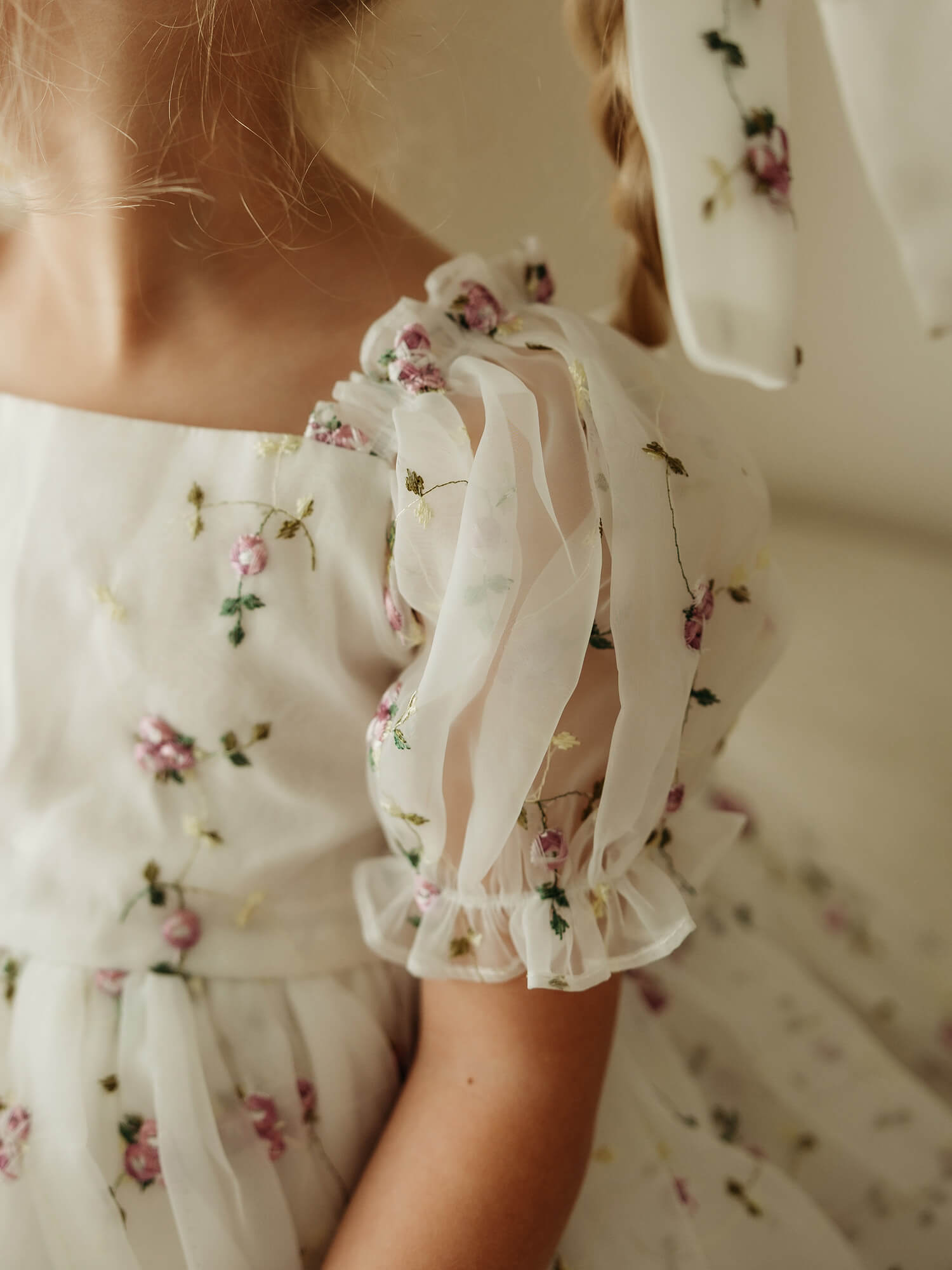 A close-up of a child wearing a white organza dress with lilac floral embroidery and puff sleeves, photographed in a softly lit indoor setting.