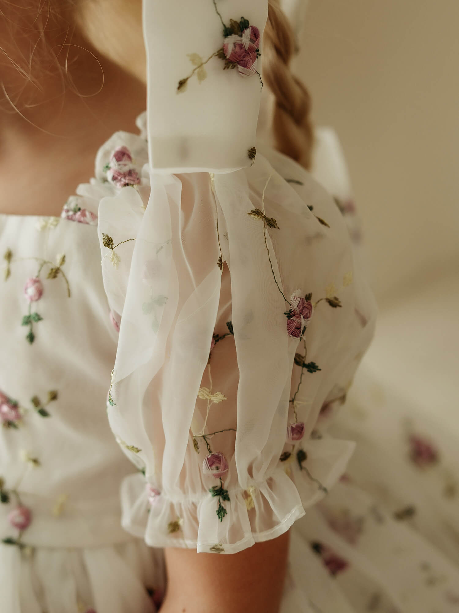 Close-up of a child wearing an organza flower girl dress with lilac floral embroidery and sheer ruffled puff sleeves, photographed in soft warm lighting.