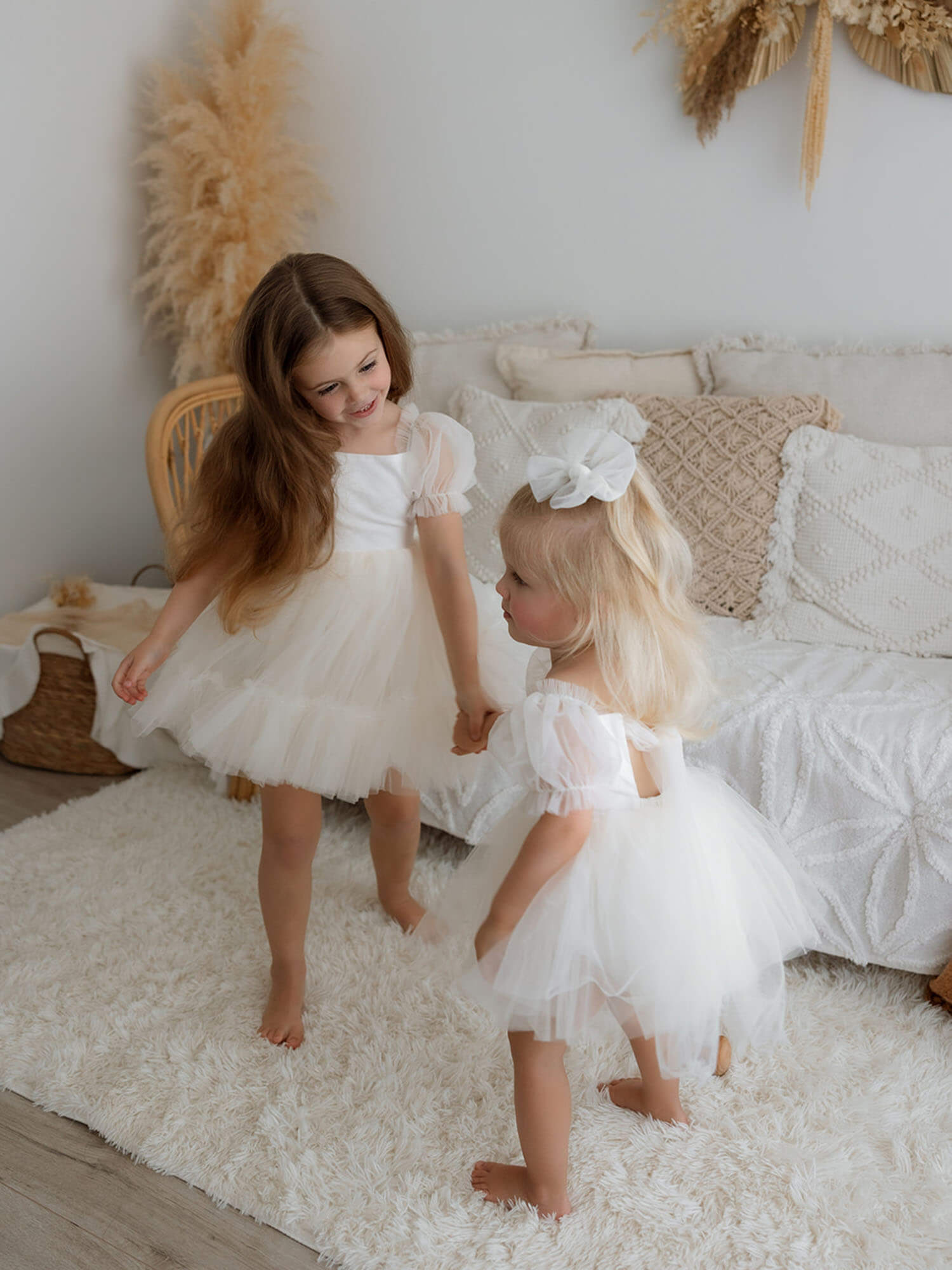 Two young sisters holding hands, one wearing a Gabrielle cream flower girl dress and the other in a Gabrielle cream baby flower girl romper with a matching tulle hair bow.