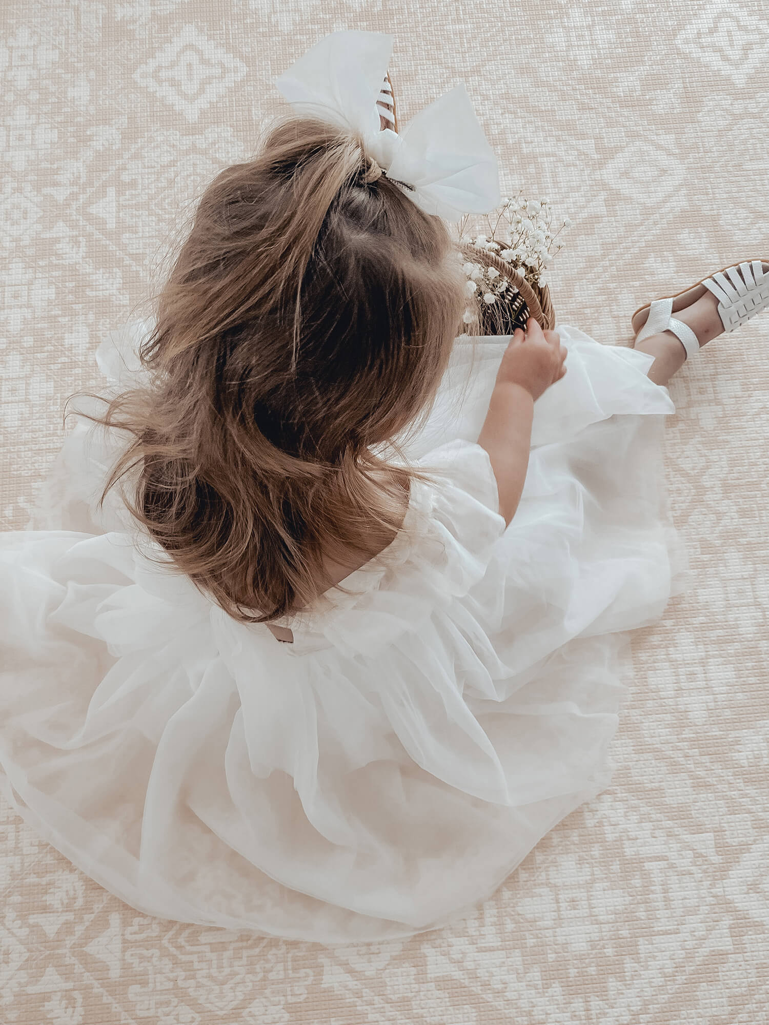 A young girl sits on the floor holding a basket of flowers, she wears our Gigi white flower girl dress.