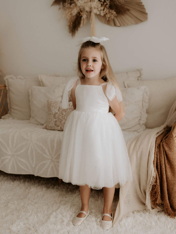 Isla ivory tulle flower girl dress is worn by a young girl. It has tulle tie sleeves and a tea length tulle skirt.