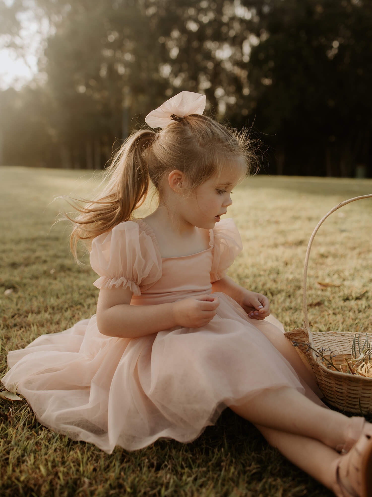 A young girl sits with a basket, she wears a tulle bow in her hair and our Champagne Layla flower girl dress.