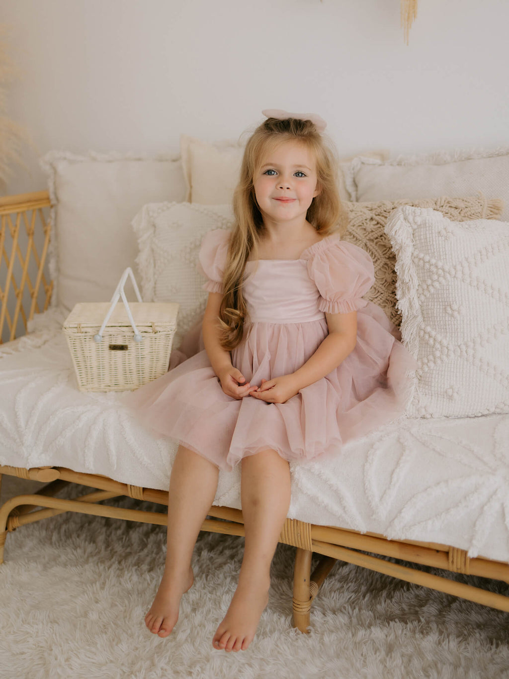 Layla puff sleeve flower girl dress and tulle bow hair clip, both in dusty pink, are worn by a young girl who sits on a rattan couch.