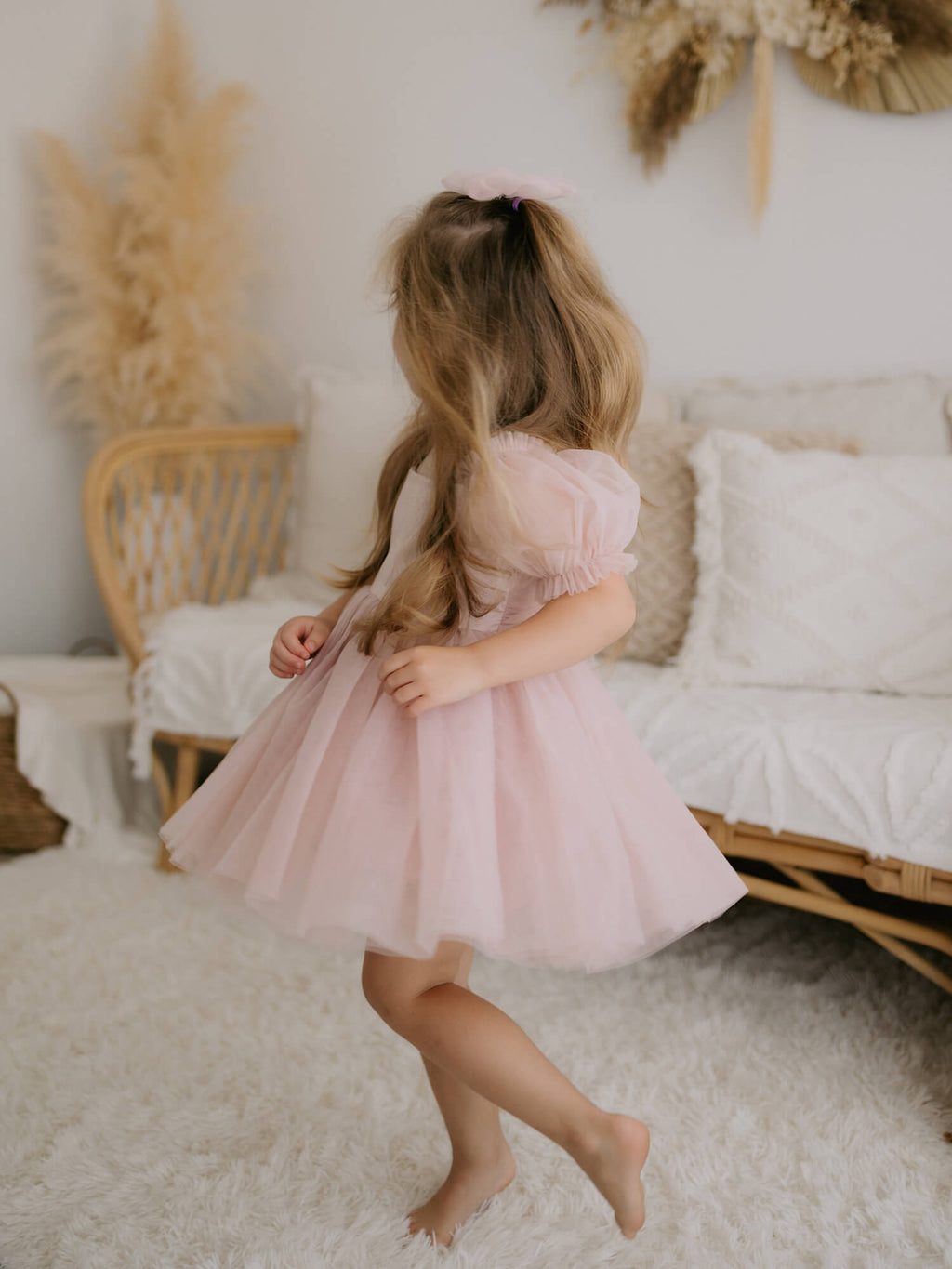 Layla puff sleeve flower girl dress and tulle bow hair clip, both in dusty pink, are worn by a young girl who sits on a rattan couch.