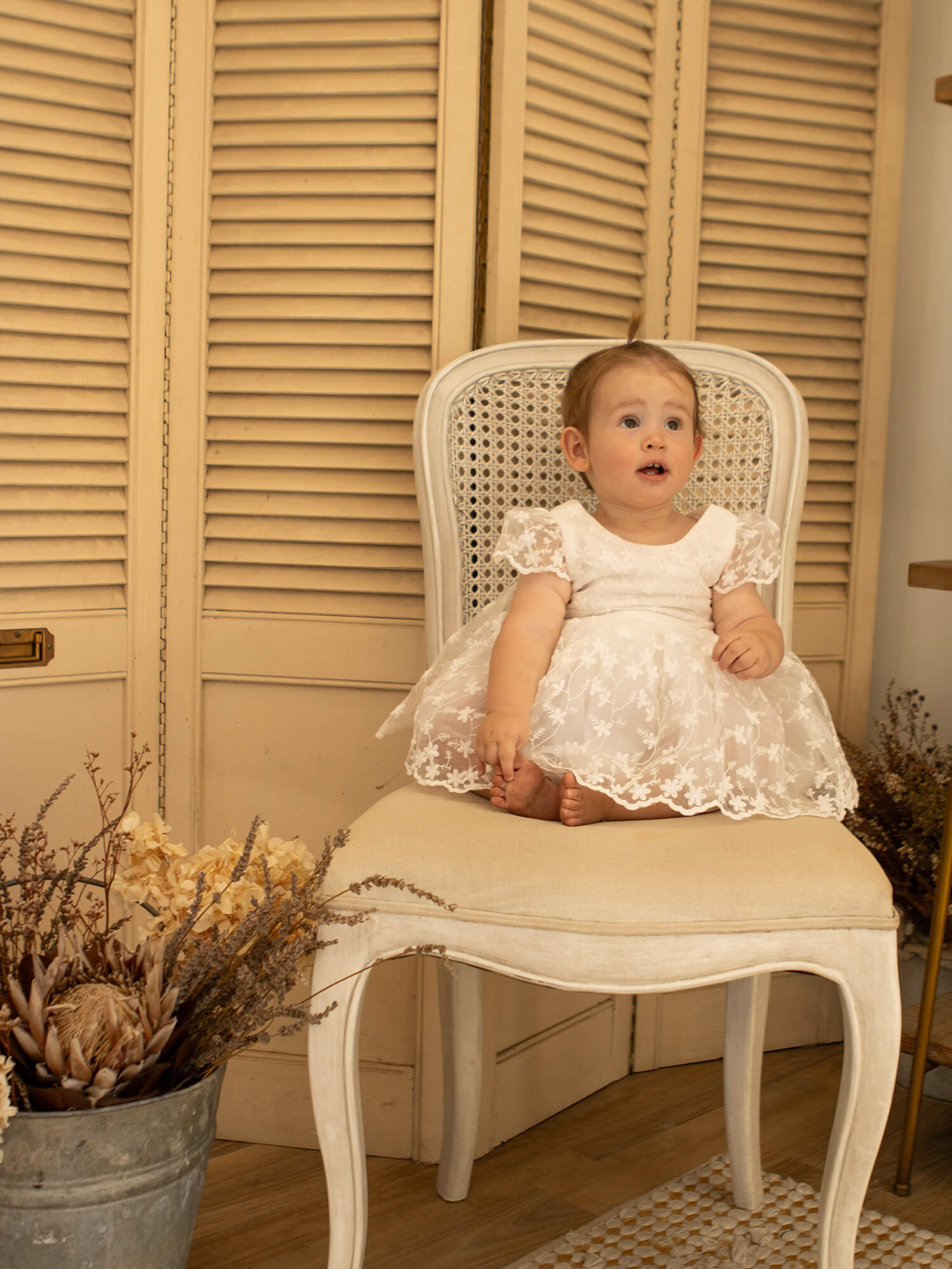 A baby girl sits wearing our Pippa baby flower girl dress in floral lace.