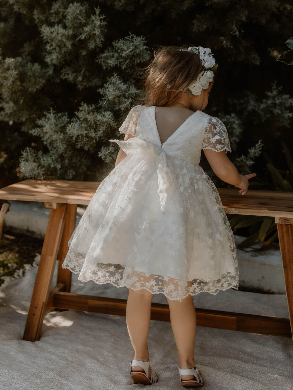 Pippa ivory floral lace flower girl dress is worn by a young girl, she sits on a stool laughing, wearing a rose flower crown in her hair.