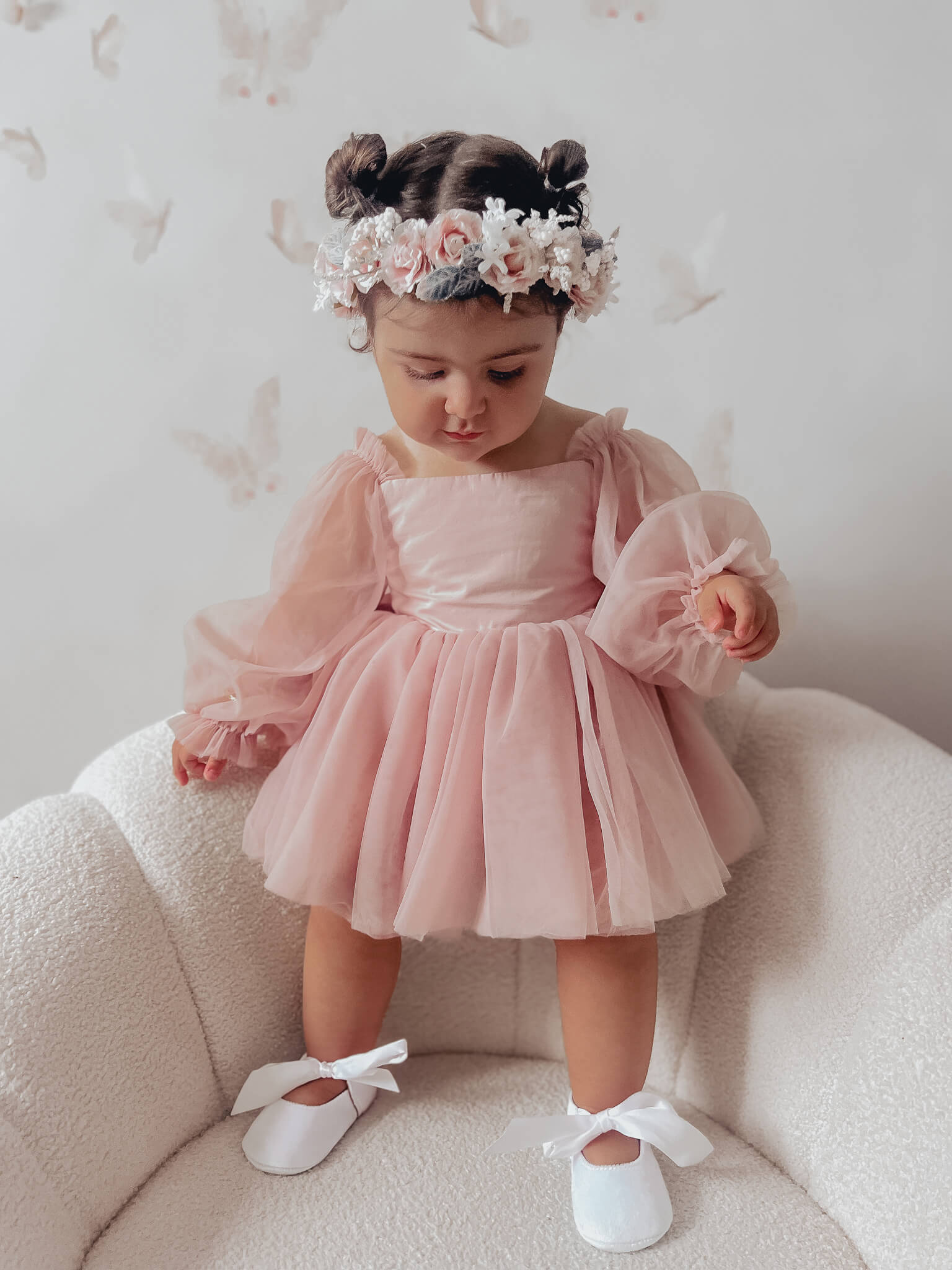 Toddler stands wearing our Wren baby flower girl romper dress in dusty pink. She wears sweet white flower girl shoes and a girls Rose blush flower crown to match the dress.