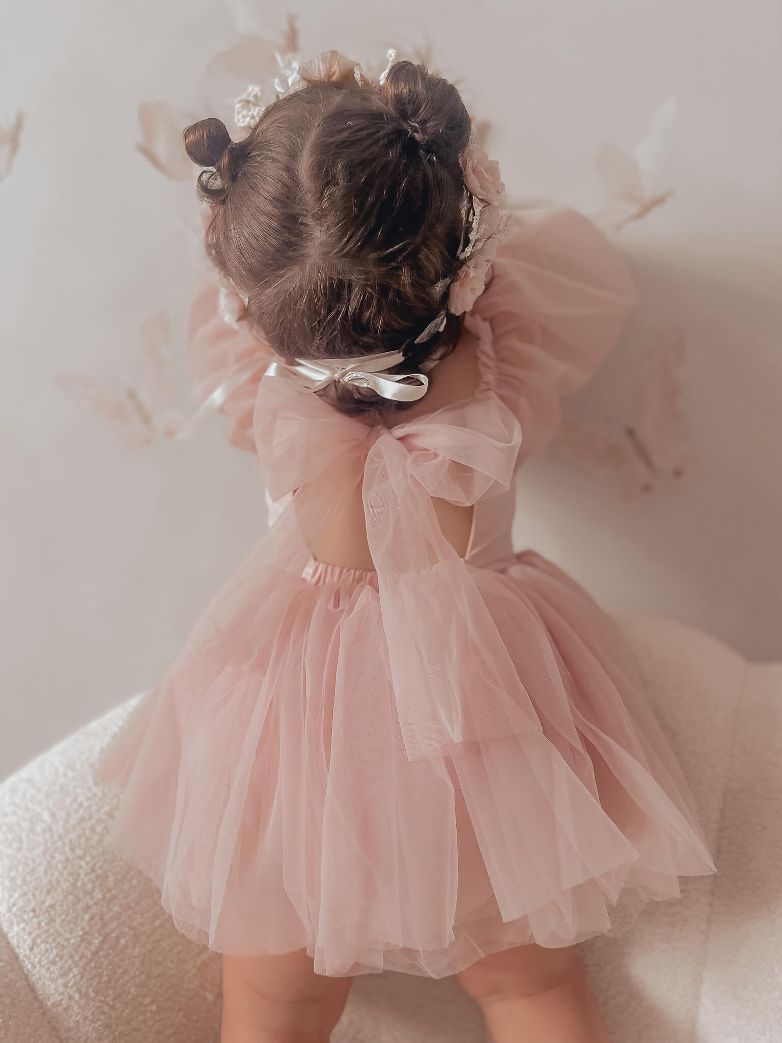 Toddler wears our Wren baby flower girl romper in dusty pink. She stands showing the back of the romper which has a large tulle bow.