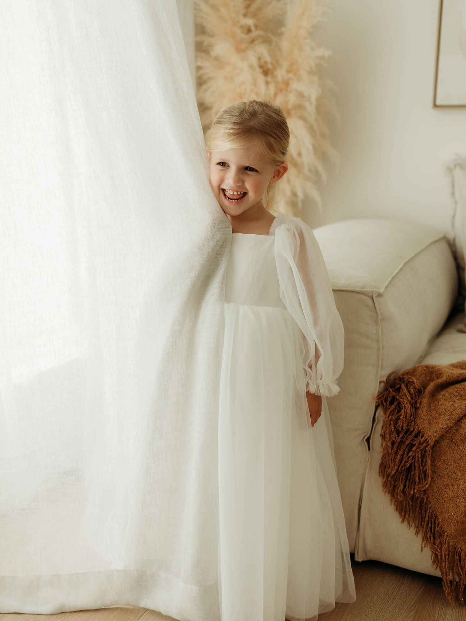 Young girl in a white flower girl dress standing in a softly lit room with neutral colours.