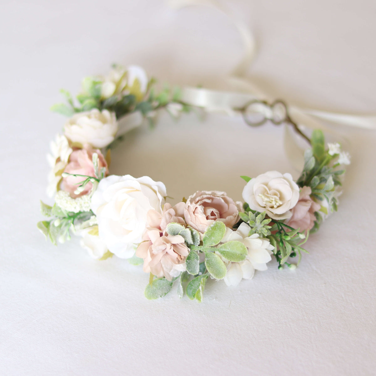 Daisy girls flower crown showing a central ivory flower, surrounded by smaller blush and ivory flowers, and green foliage. A tie back floral wreath with an ivory ribbon.