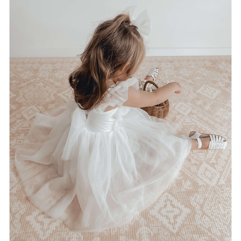 Gigi white flower girl dress worn by a young girl, she also wears our ivory tulle bow in her hair and stands beside a basket of flowers.