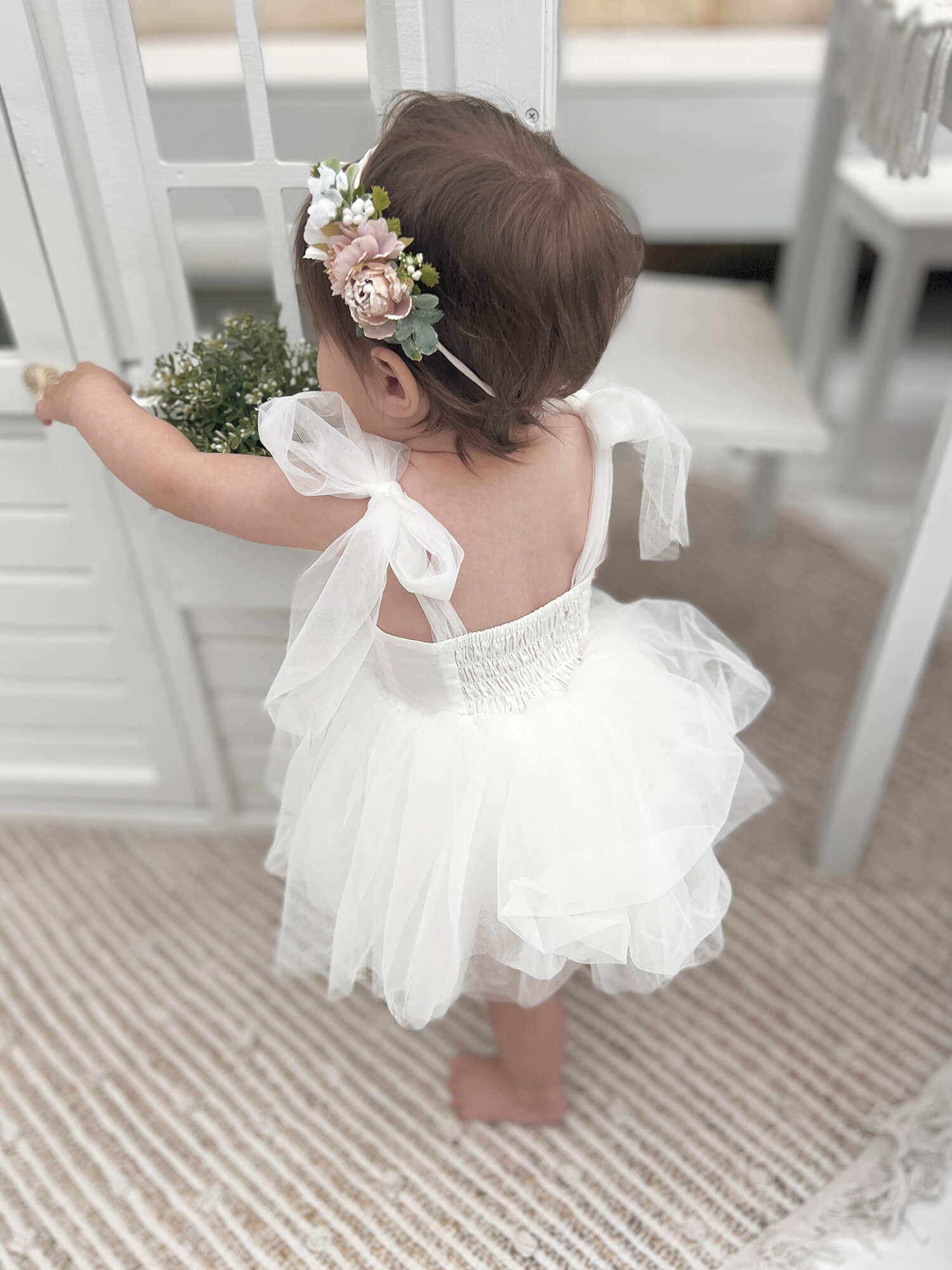 A baby stands wearing our Harper baby flower girl romper dress, with shirred back and tulle tie straps. She also wears our Bryony dusty pink baby floral headband.