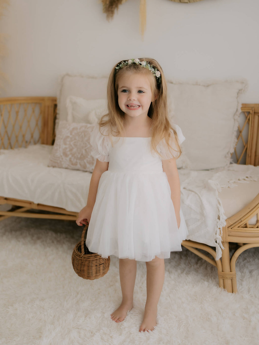 Layla flower girl dress in ivory is worn by a young girl. She stands holding a basket and smiling. She also wears our Eden flower crown hair wreath.