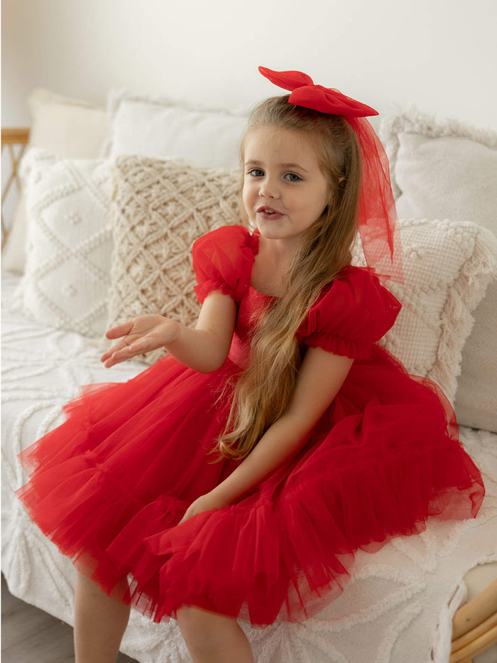 Gabrielle ruby red puff sleeve Christmas dress is worn by a young girl, she also wears a large red tulle bow in her hair.