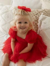 A toddler sits smiling, she wears a red tulle bow in her hair and a red romper dress with tulle puff sleeves, our  Gabrielle romper in ruby red.