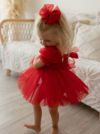 A toddler twirls wearing our Red toddler Christmas dress, Gabrielle romper in ruby red. She also wears a medium red tulle bow in her hair.