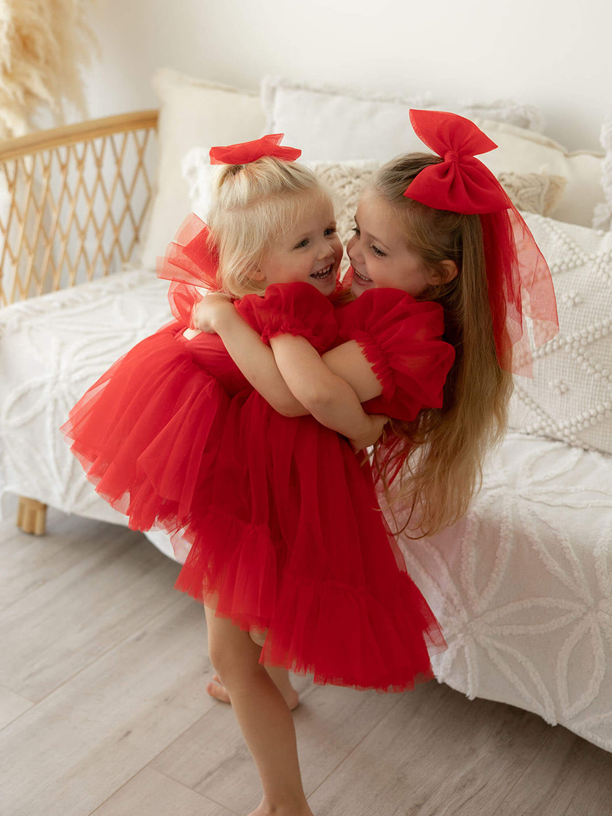 A big sister holds her little sister, both wearing matching red tulle Christmas dresses and tulle hair bows.