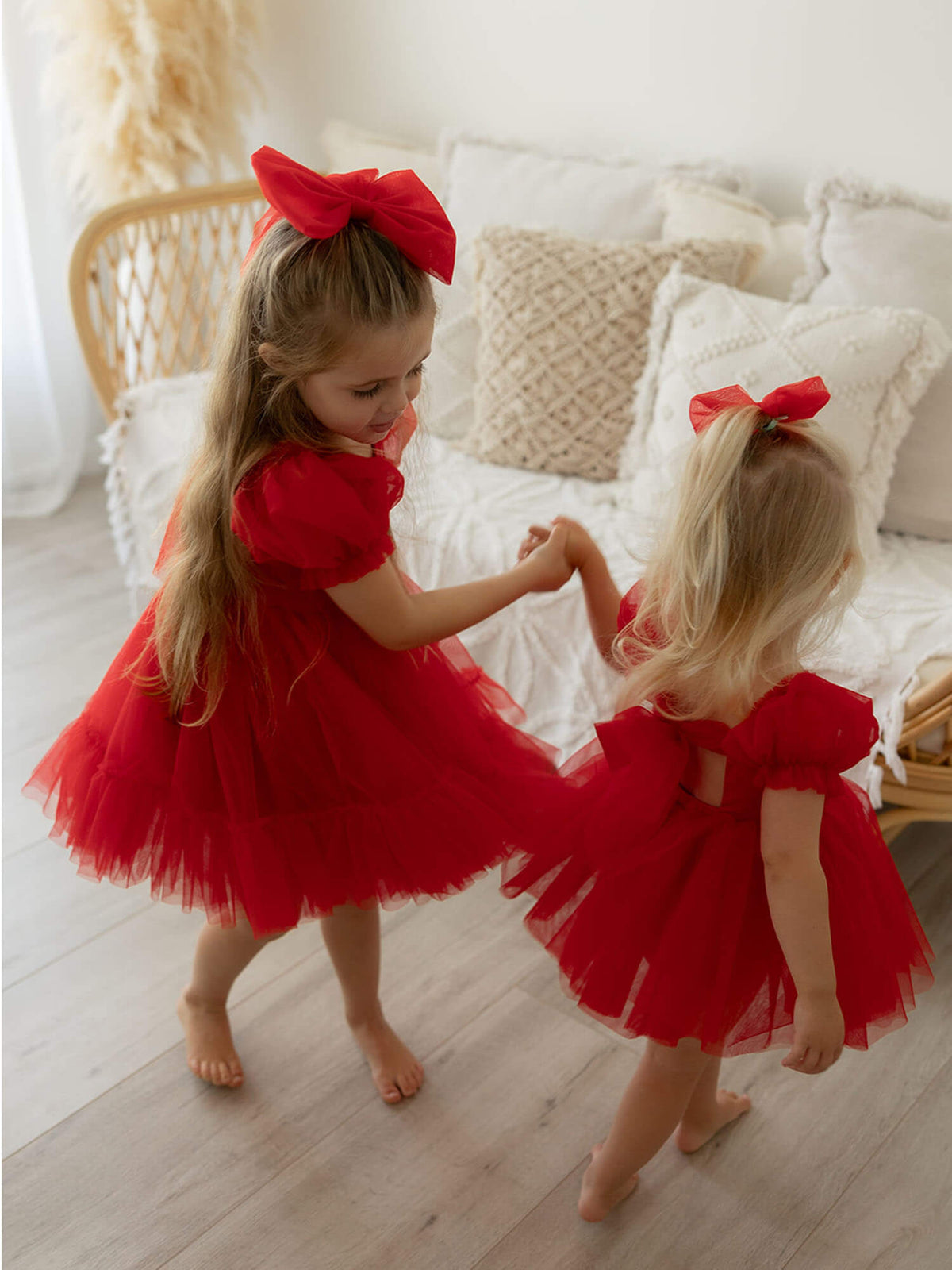 Sisters hold hands dancing wearing matching red girls dresses and matching red tulle bows.