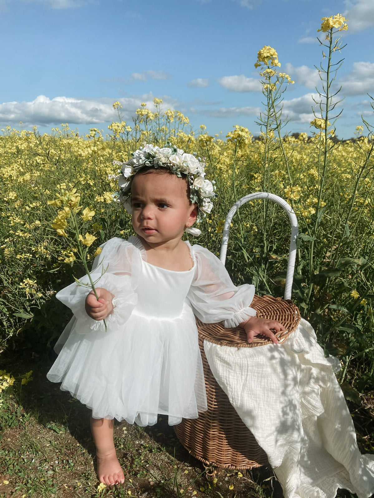 Eva baby flower girl romper is worn by a toddler standing in a flower field. She also wears our Anais flower crown.