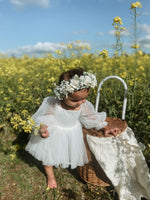 A toddler looks at a picnic basket, wearing our Eva baby flower girl romper with full length tulle sleeves, in ivory. She stands in a flower field surrounded by yellow flowers.